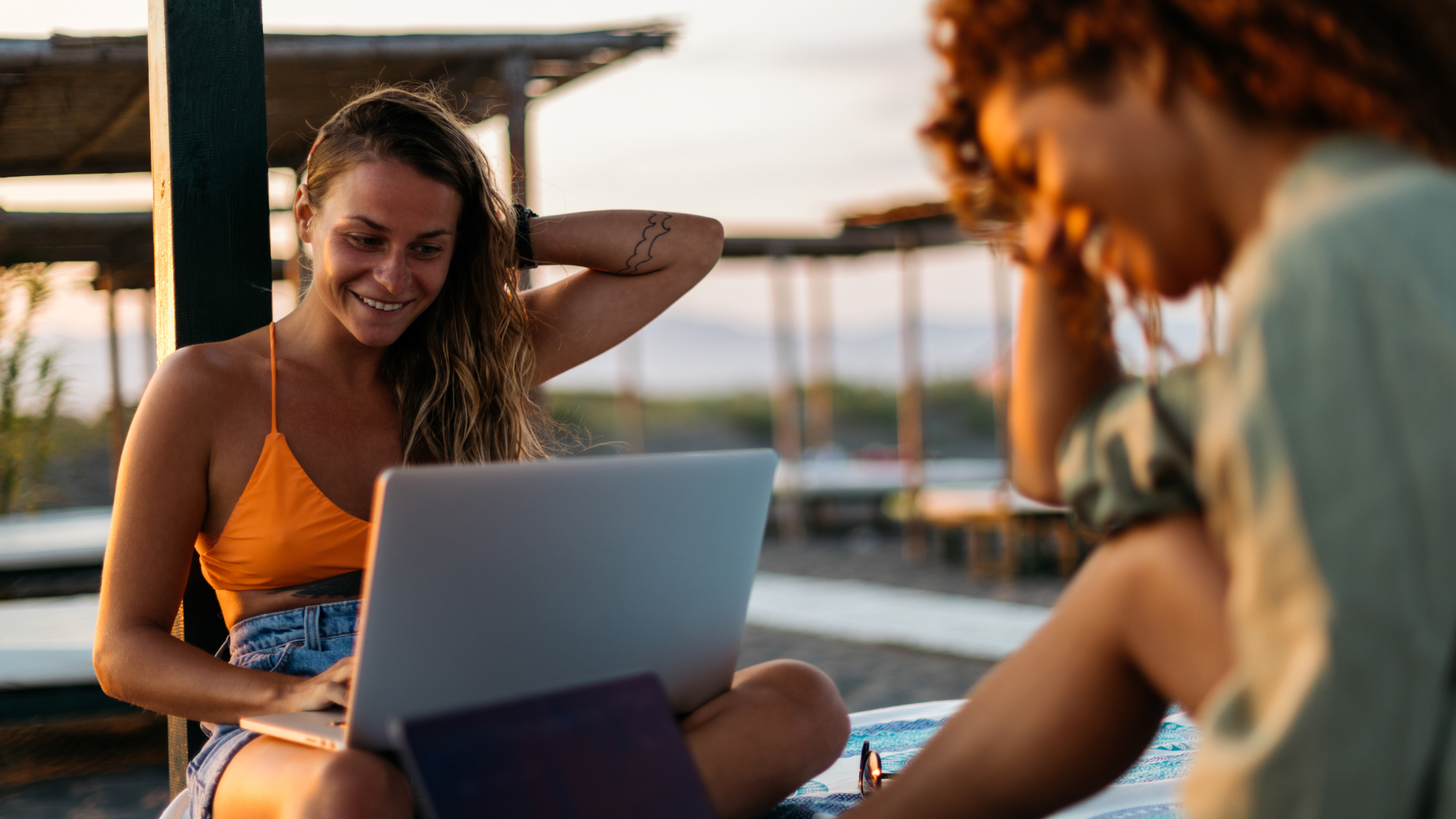 two women sitting at a beach with a laptop